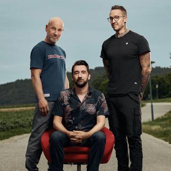 Roland Düringer stehend, Bernhard Egger sitzend auf einem roten Stuhl und Bernhard Egger stehend rechts davon.